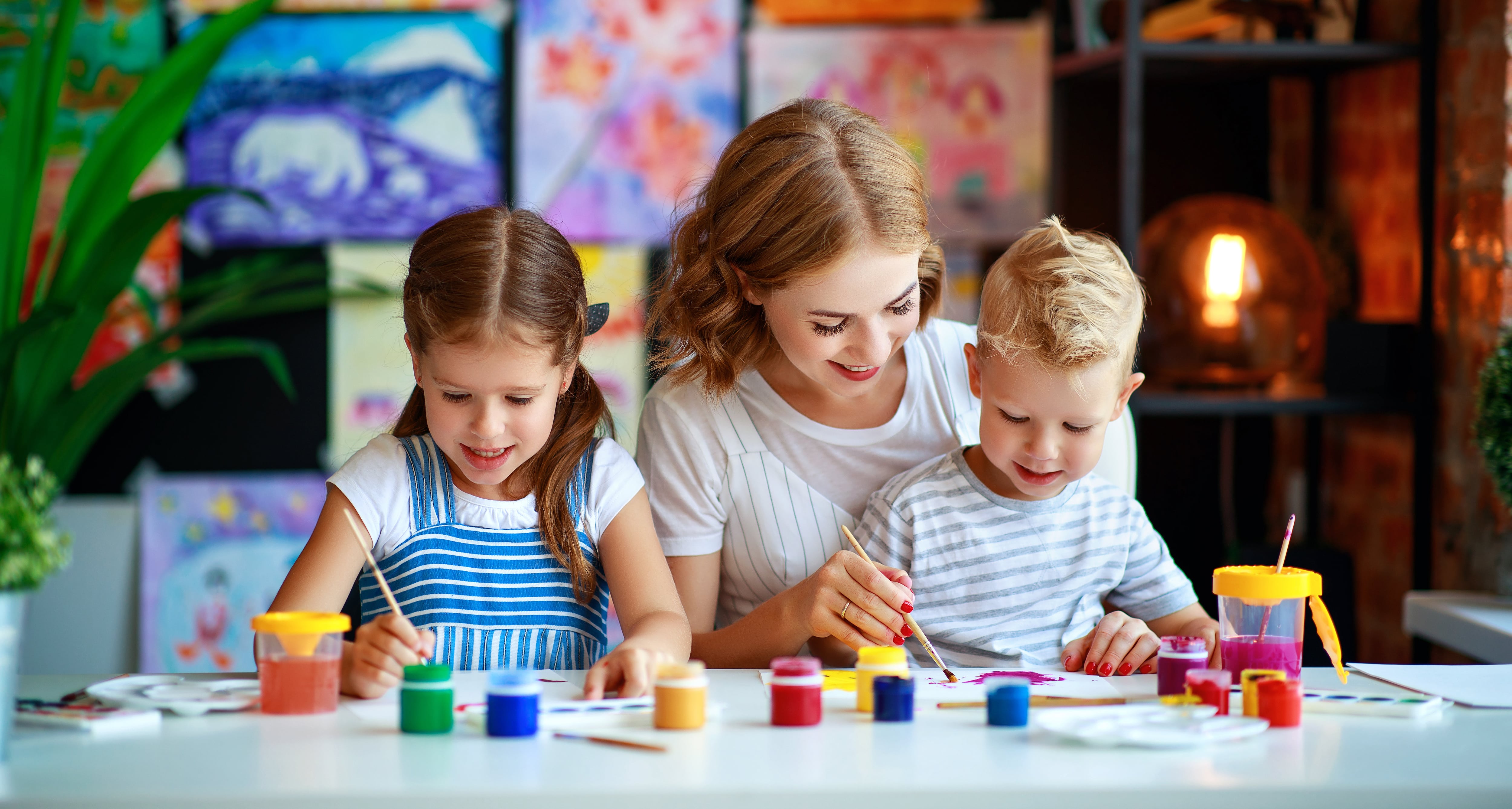 A mom painting with her kids with paintings from the kids hung up behind them