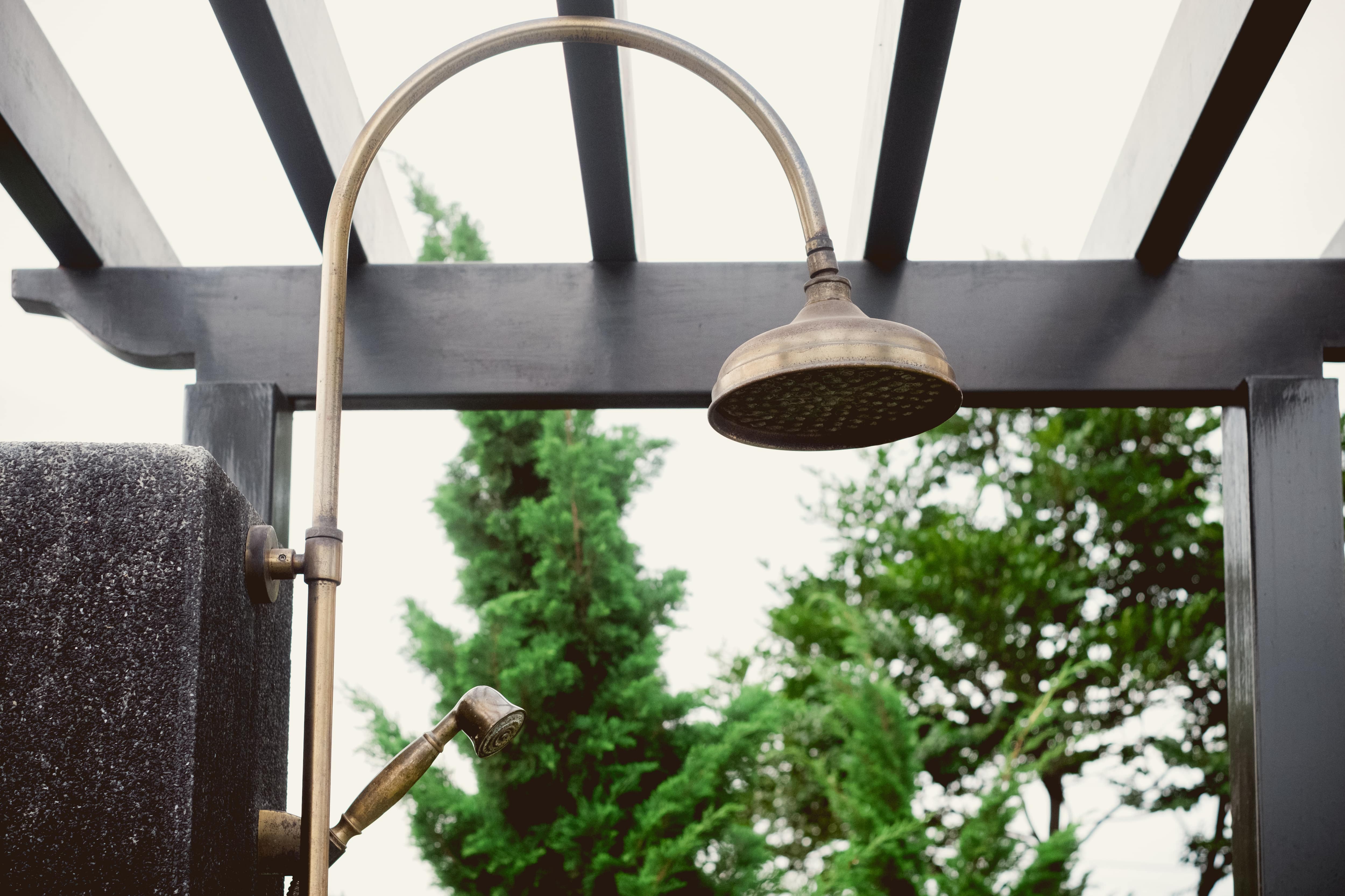 An arched brass showerhead outdoors underneath a wooden trellis 