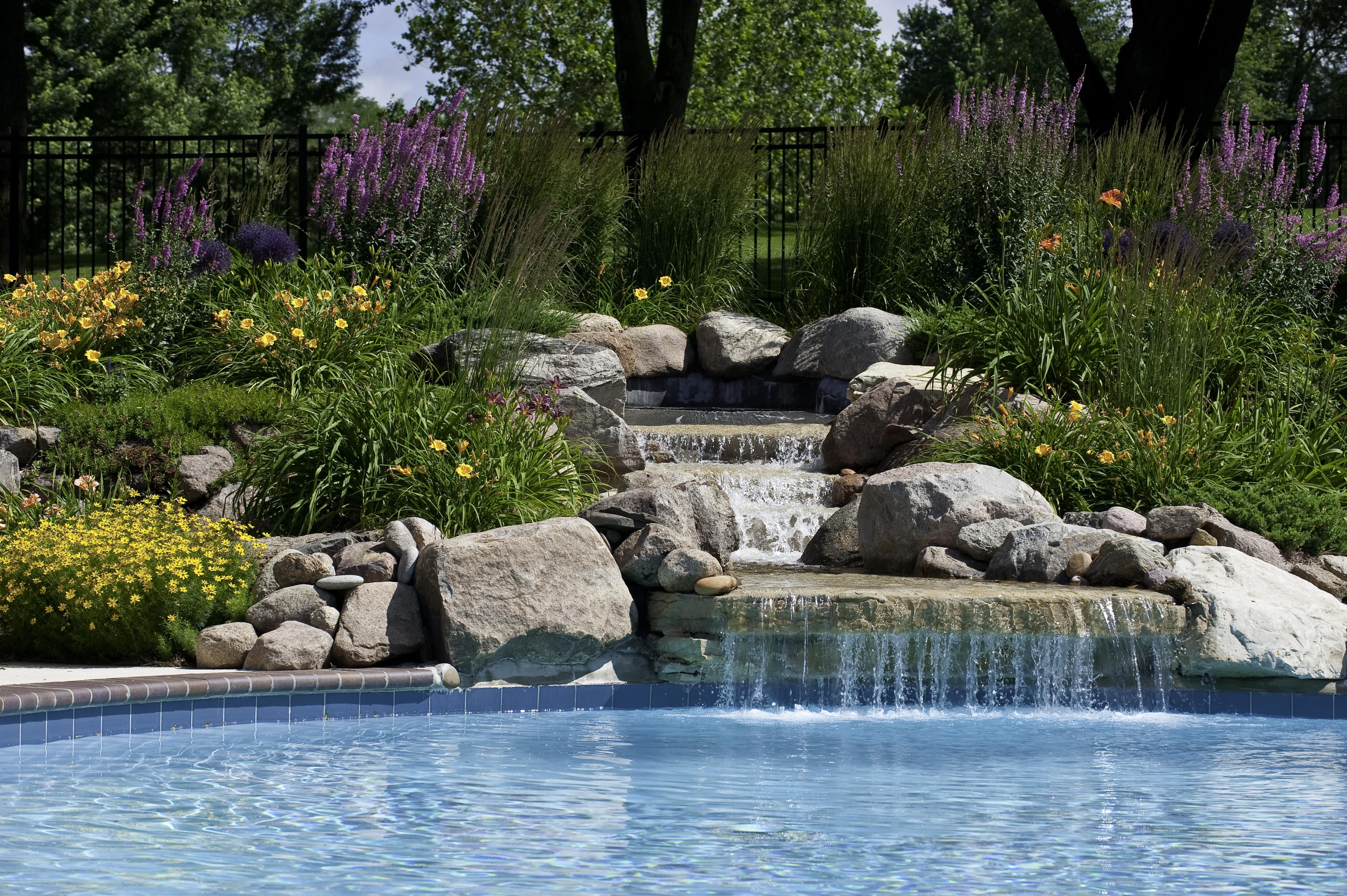 A waterfall built with rocks emptying into a pool surrounded by flowers and greenery 