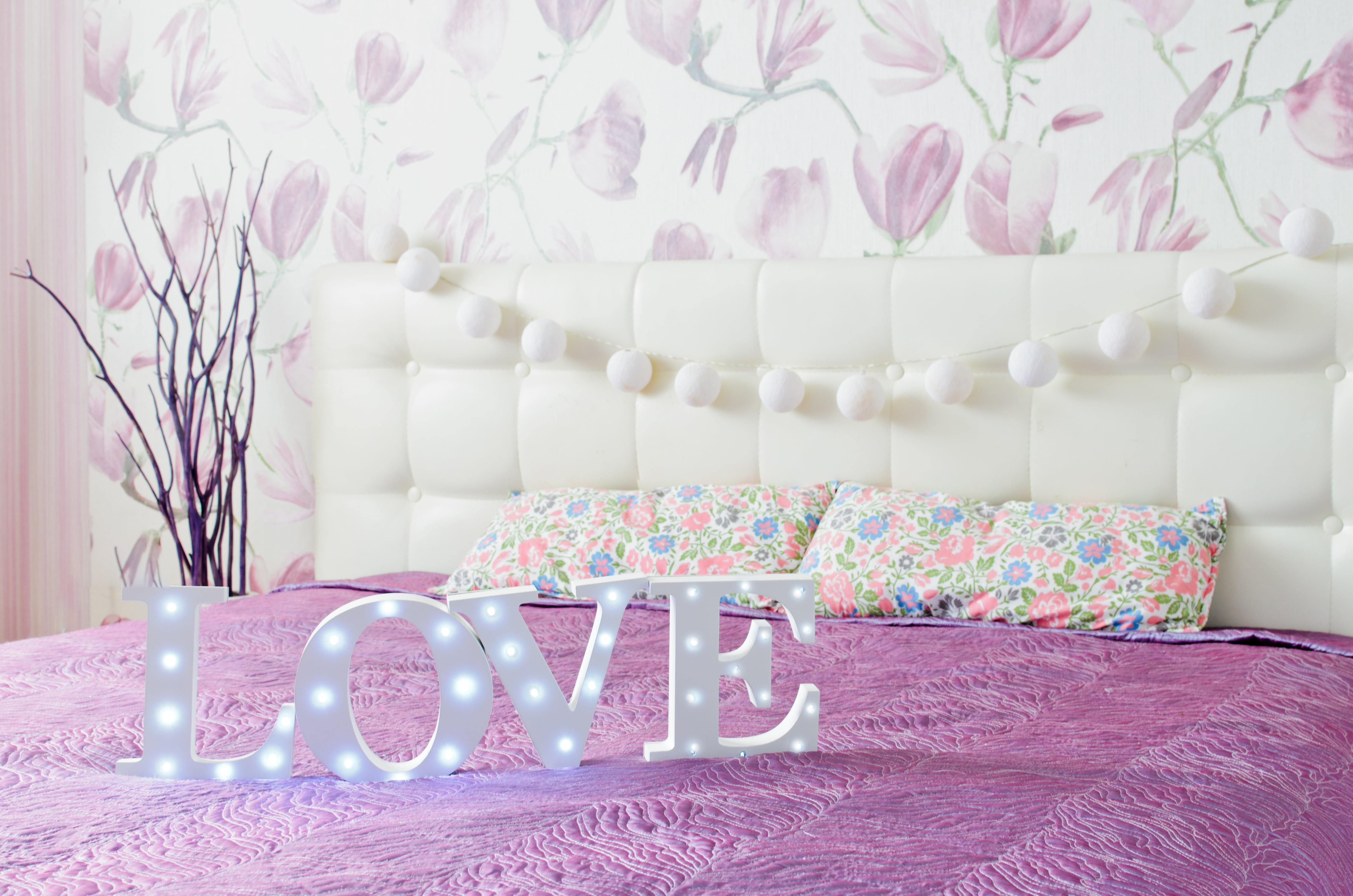 Bedroom with purple flower wallpaper, white ball garland on white tufted headboard, and Love light-up sign on purple bedding 