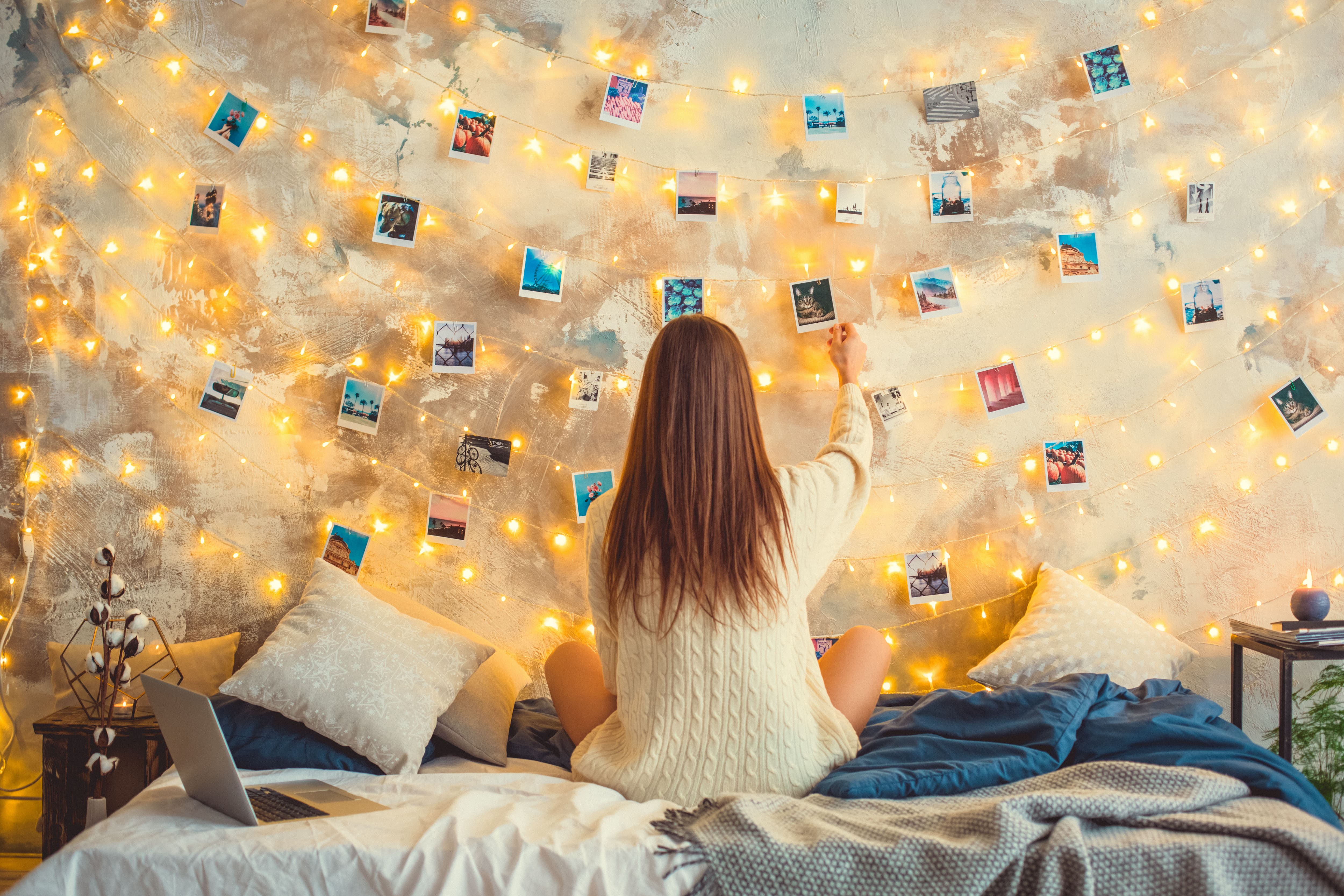 Teen girl sitting on bed while decorating the wall behind it with photos and string lights 